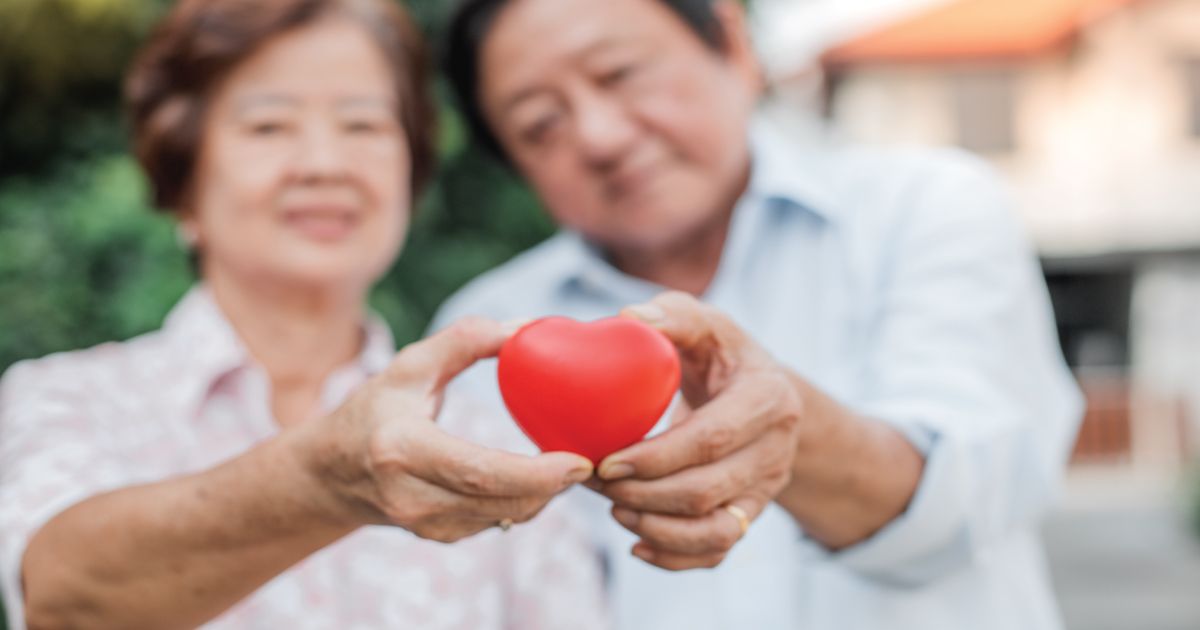 Hands holding a small red heart in the foreground, with a smiling couple blurred behind