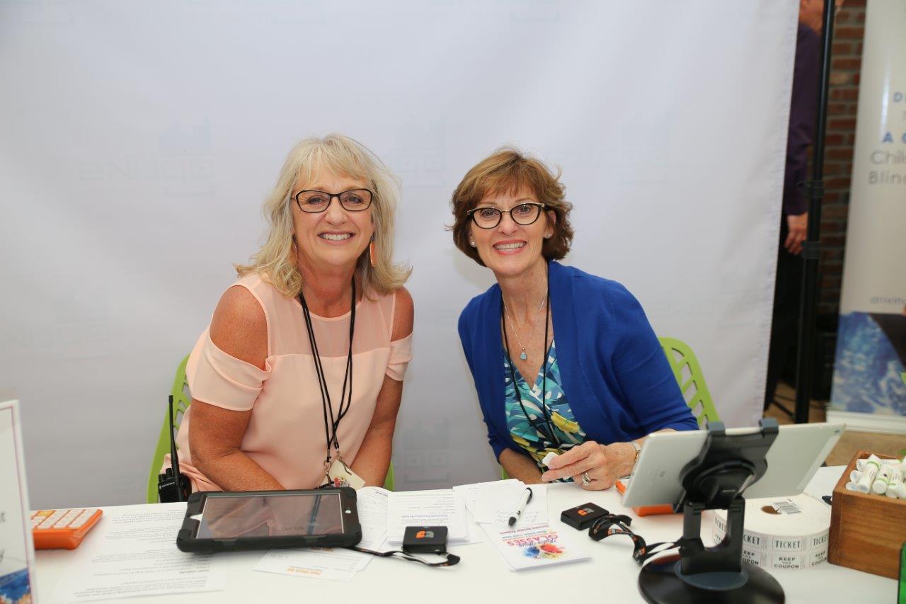 Two smiling people wearing glasses reception table at 2018 Raise Your Glasses