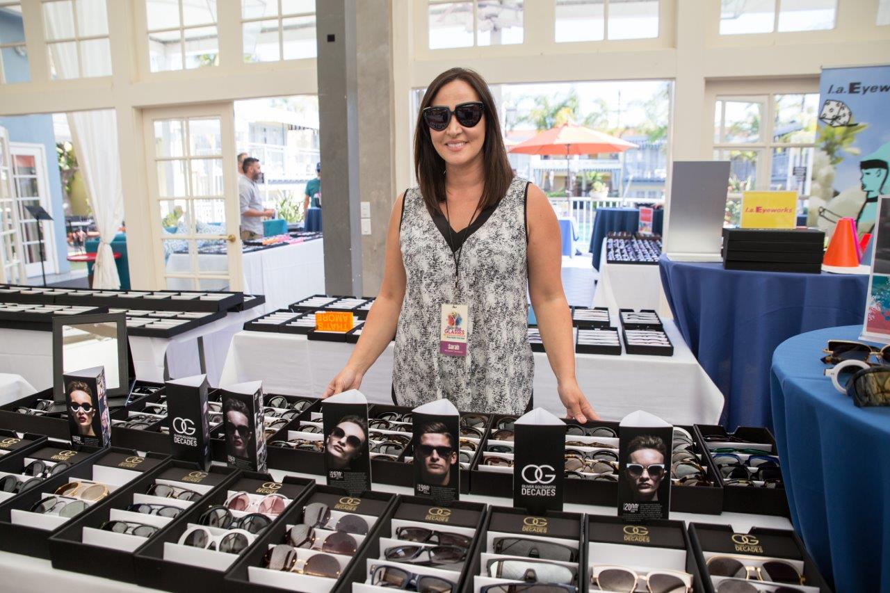 Vendor standing behind tabletop display of Oliver Goldsmith eyewear at 2018 Raise Your Glasses