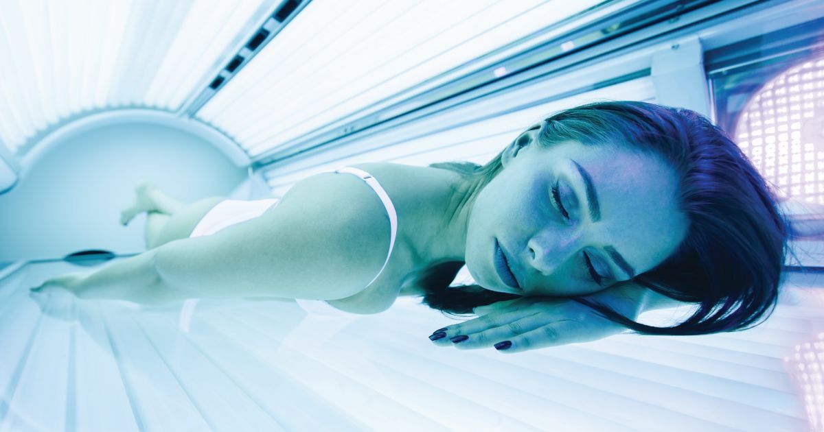 Woman lying inside an indoor tanning bed illuminated by blue UV light with eyes closed during a tann