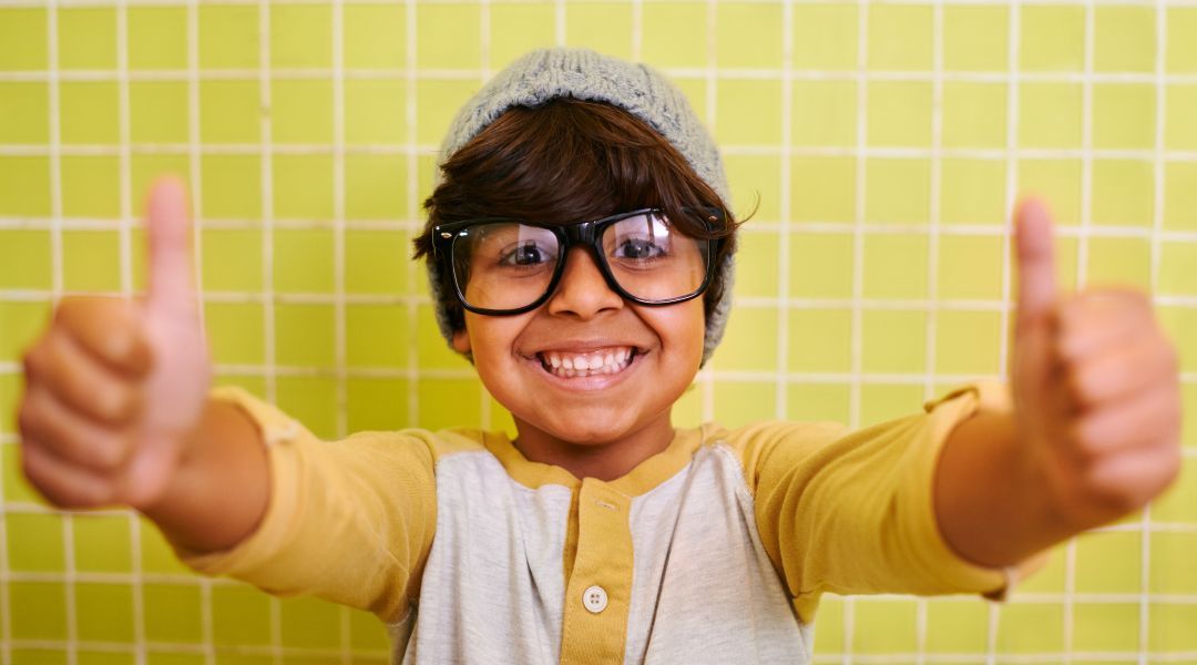A young boy wearing glasses and a hat giving two thumbs up.