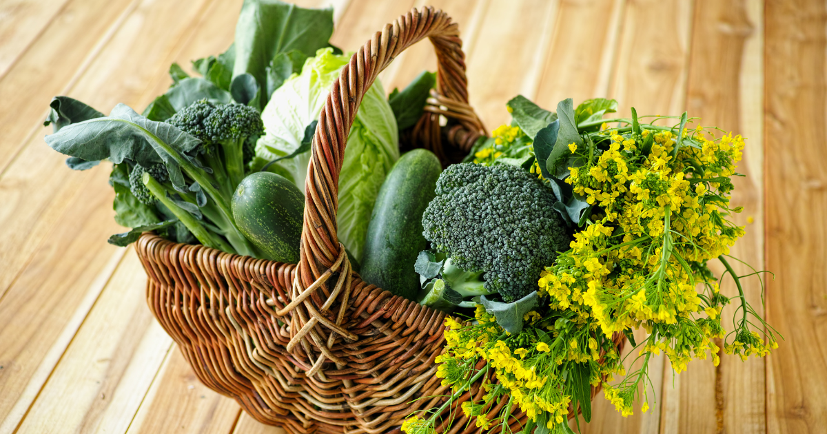 A wicker basket filled with green vegetables and yellow flowers on a wooden table .