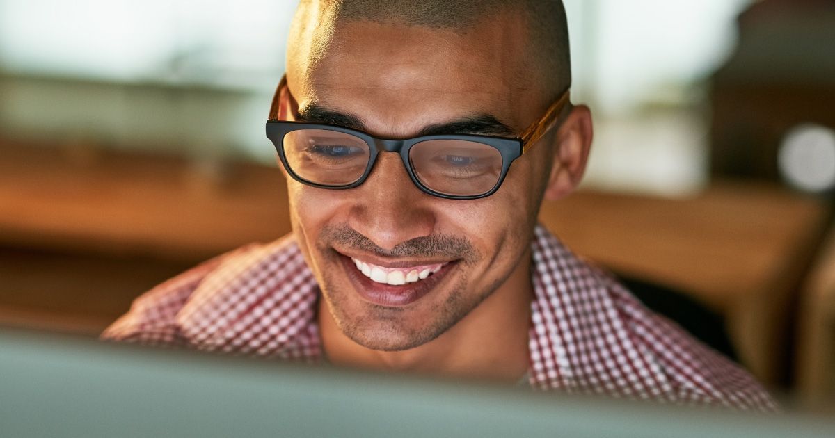 Man with glasses smiling, looking at an illuminated computer screen