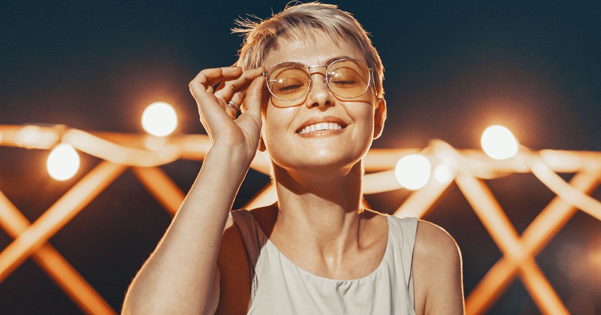 A smiling woman with her eyes closed is sitting in front of a railing with light bulbs.