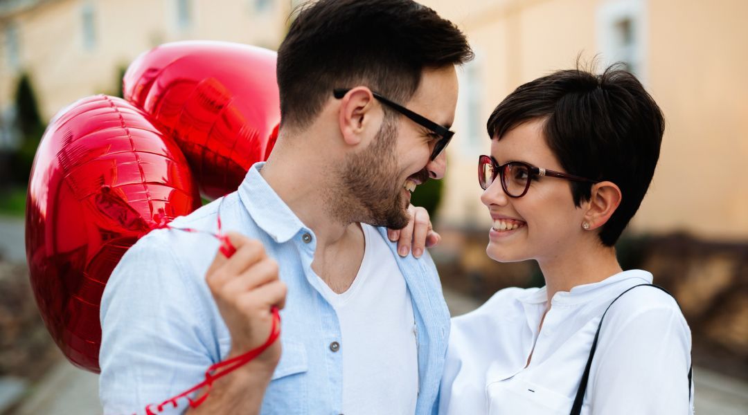 A man and a woman are looking at each other while holding red heart shaped balloons .