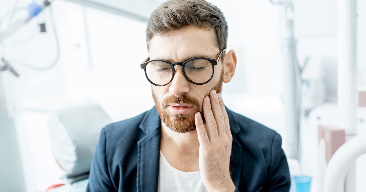 A man with glasses is holding his cheek from tooth pain.