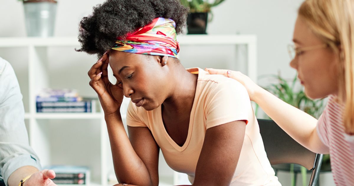 A woman is comforting another woman who is sitting with her head in her hands.