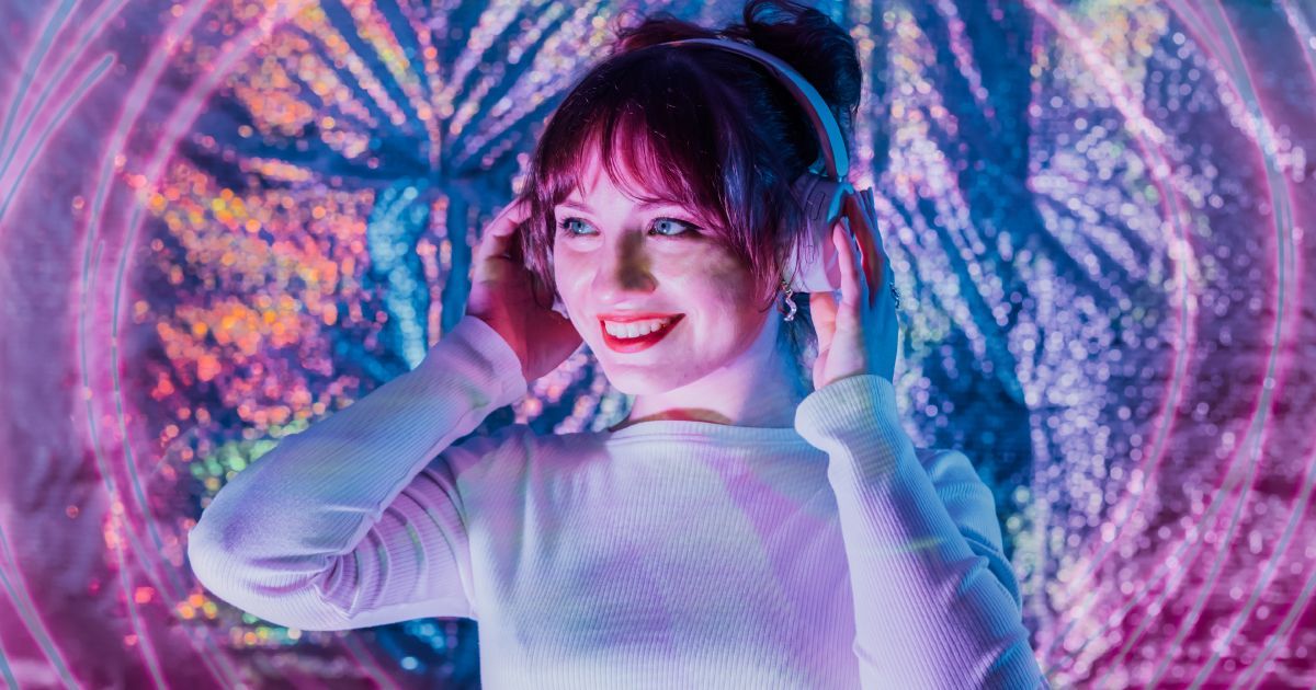 A smiling woman wearing headphones is standing in front of a colorful background.