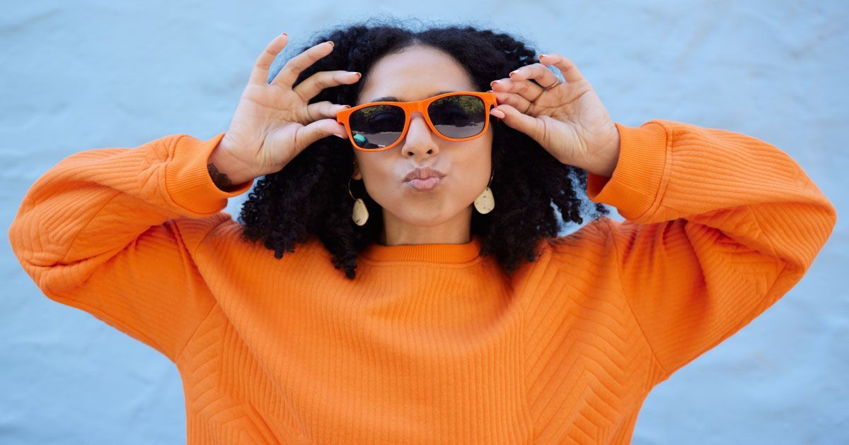 A woman wearing an orange sweater and sunglasses is blowing a kiss.