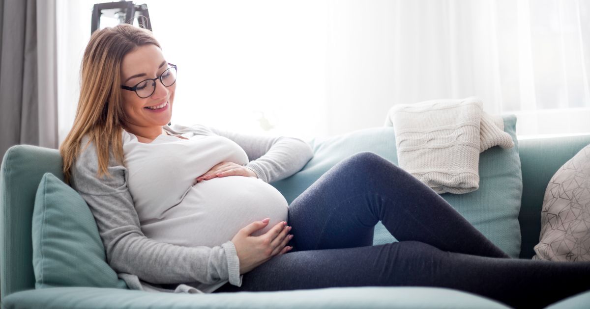 A pregnant woman wearing glasses is sitting on a couch holding her stomach.