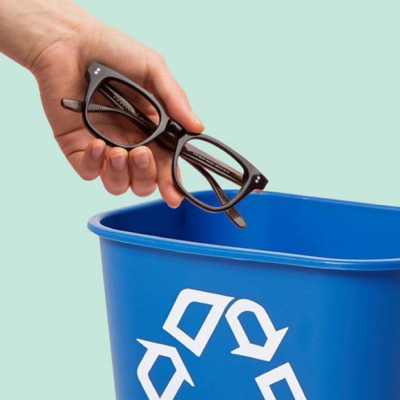 A hand is holding a pair of glasses over a blue recycling bin