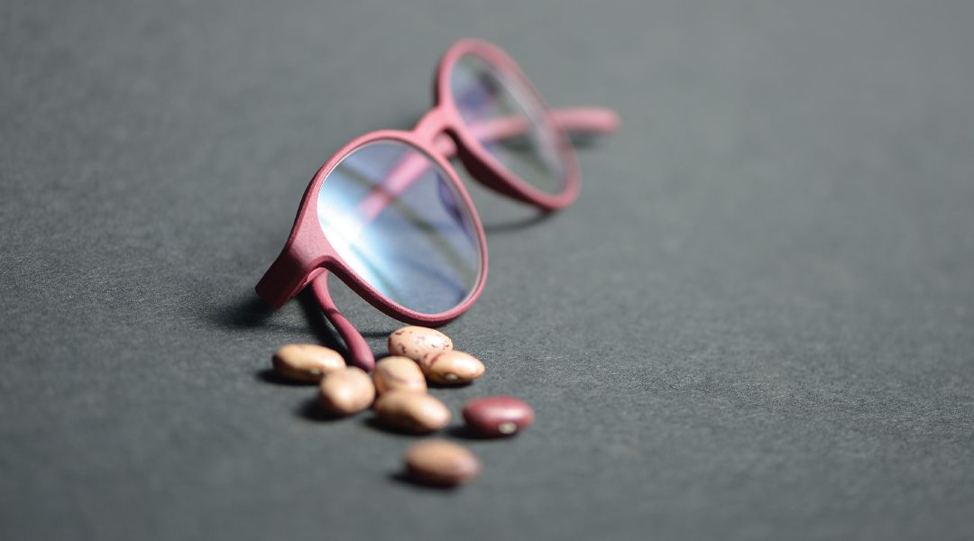 Pair of burgundy ROLF Substnce glasses siting next to several caster beans on a piece of slat