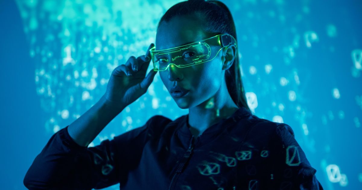 A woman standing in front a scifi background is wearing futuristic glasses.