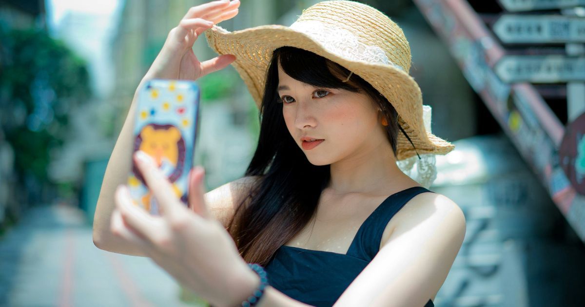 A woman wearing a straw hat is taking a selfie with her phone.