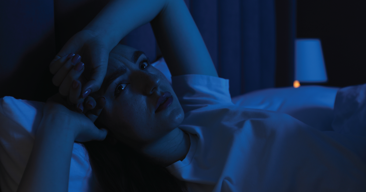 Person lying in bed at night, lit by blue light, staring upward with hands near their face.