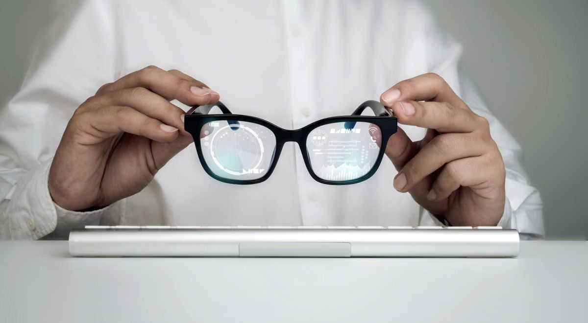 A person is holding a pair of smart glasses above a closed laptop on a desk.