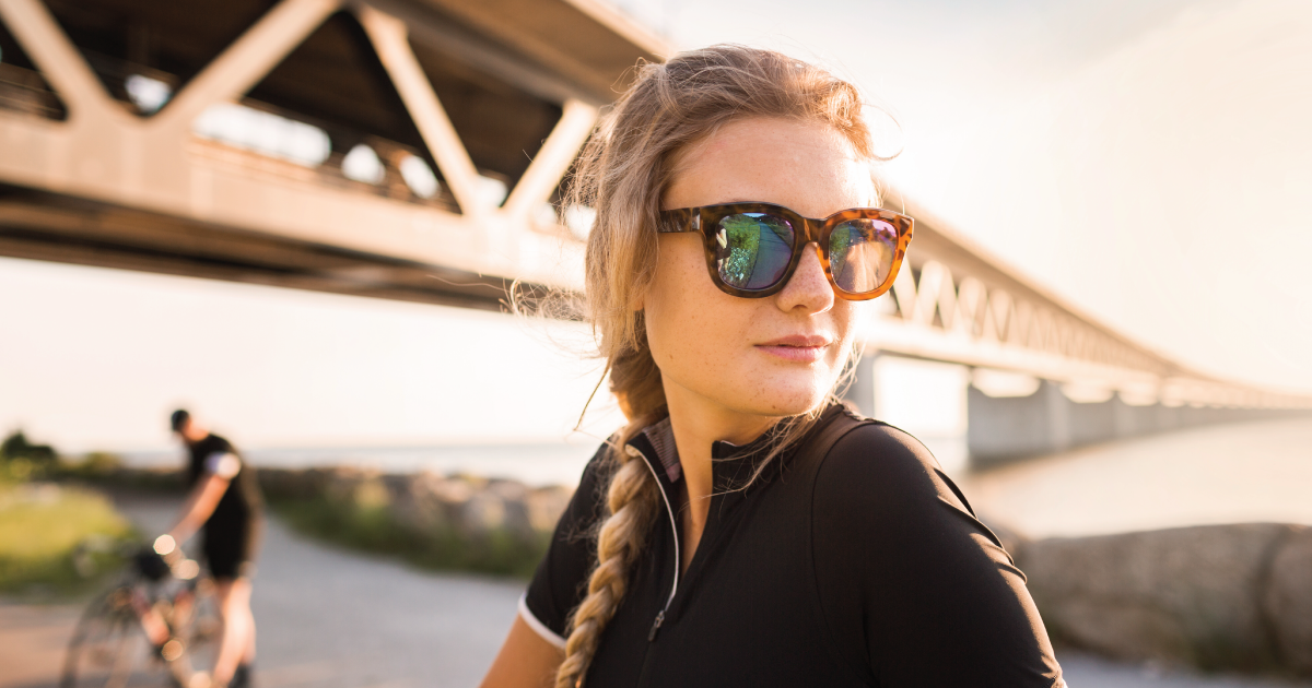 Woman in sunglasses standing under a bridge near water, with a blurred cyclist in the background.