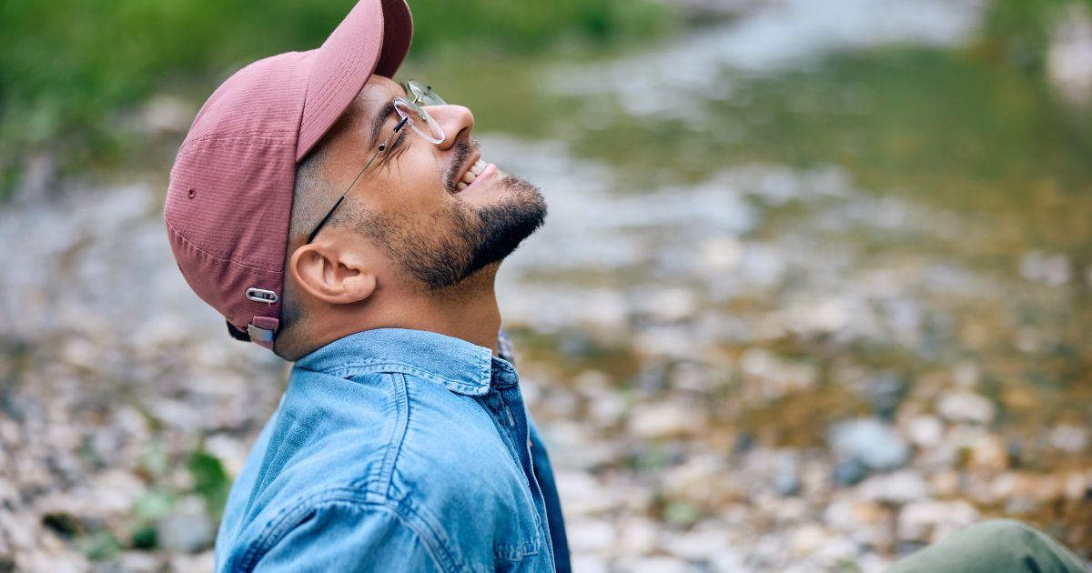 A man wearing a baseball cap and glasses is sitting next to a river looking up.