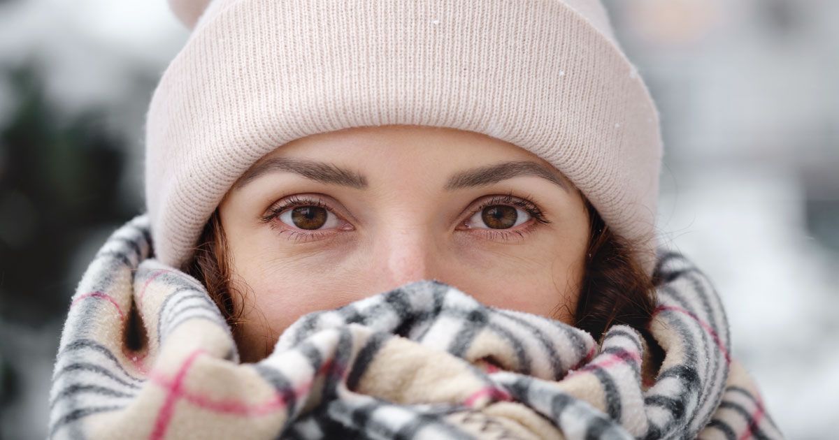 A close up of a woman outside in the snow wearing a hat and covering her face with a scarf.