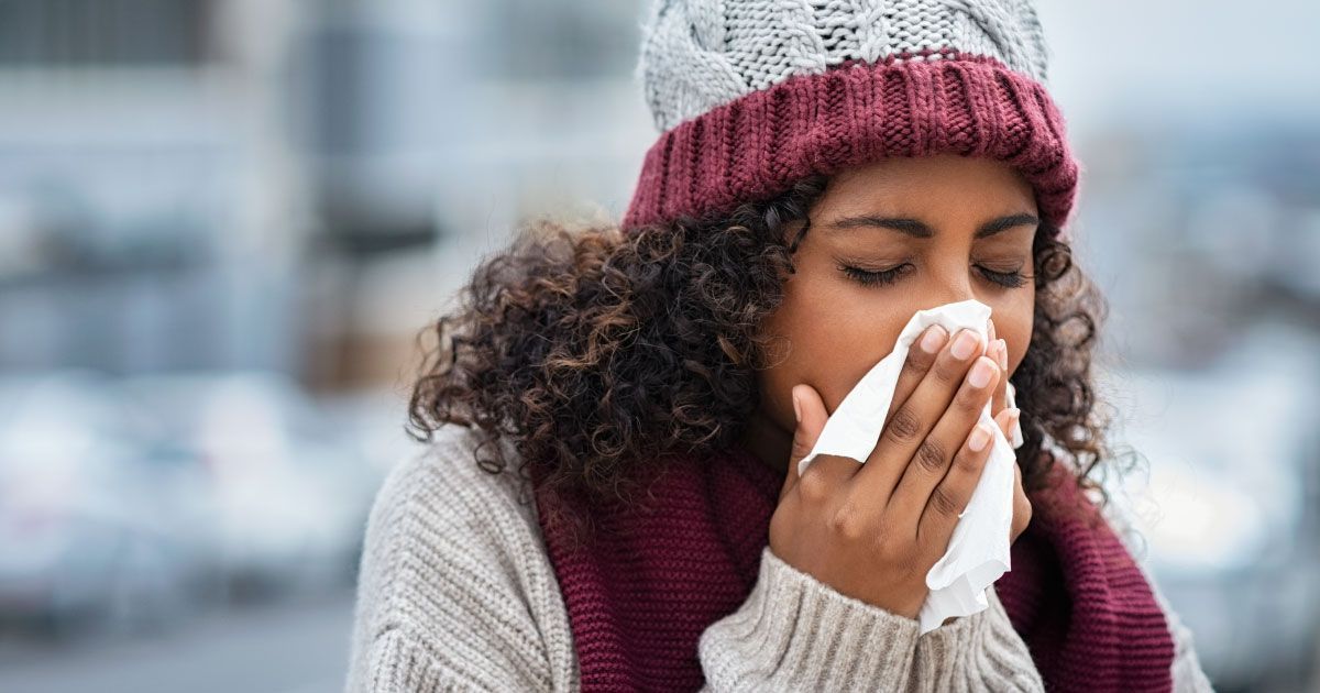 A woman wearing a hat and scarf is blowing her nose into a napkin.