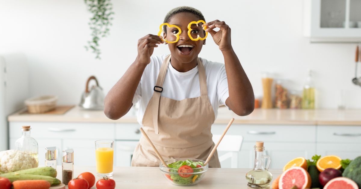 A woman in a kitchen holding vegetables in front of her eyes which looks like glasses.