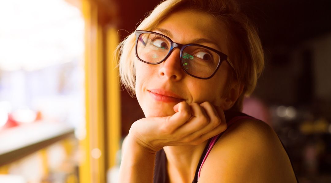 A woman wearing glasses with her hand on her chin sitting in front of a window 