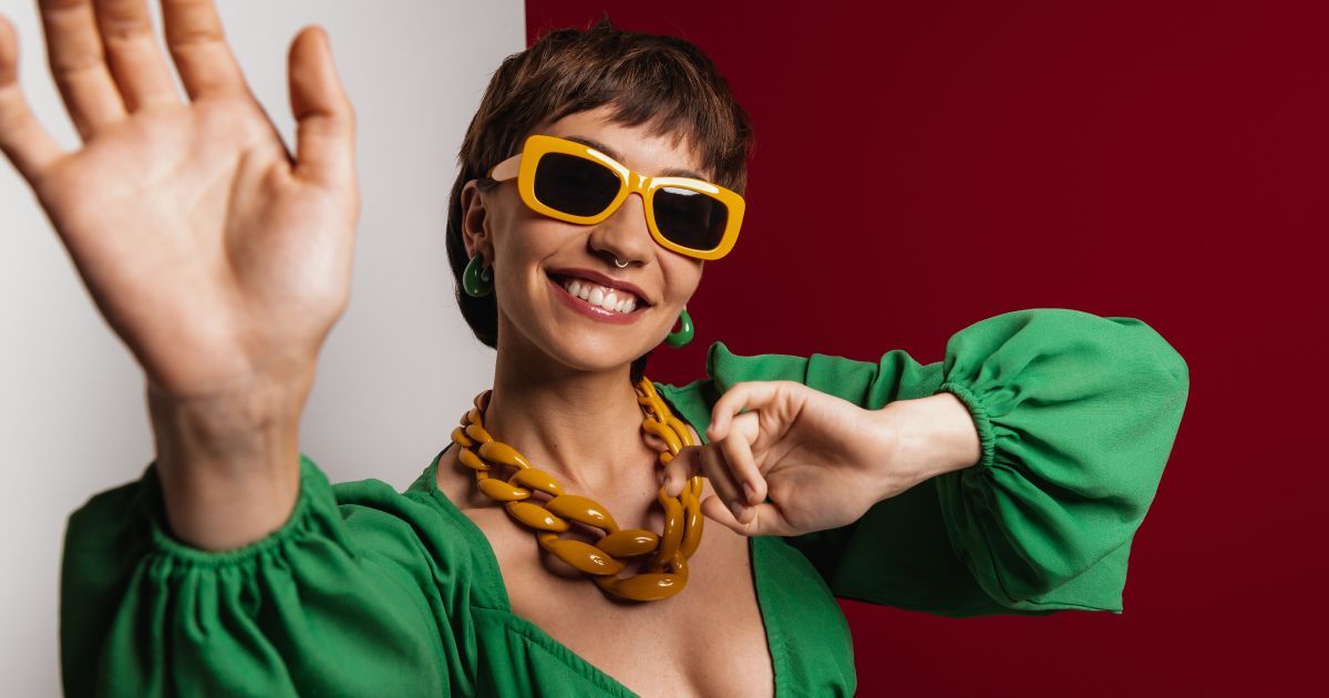 A happy woman wearing a green dress and earings with yellow sunglasses and necklace waving her hand.