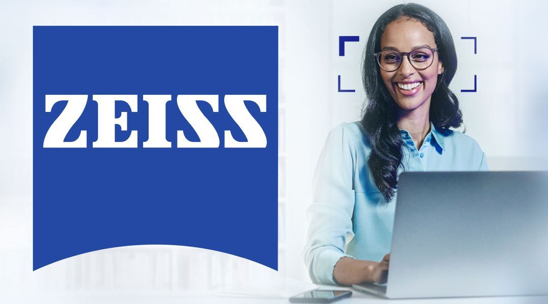 Woman in glasses sitting at a desk with a laptop next to a large ZEISS logo