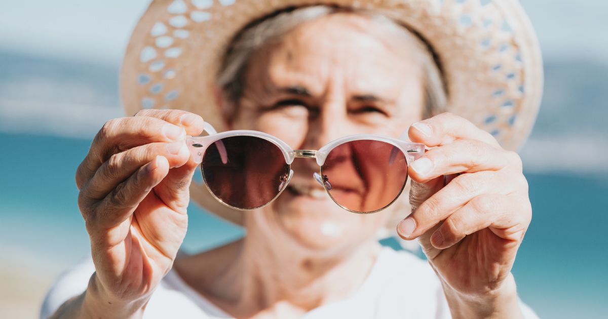 Woman holding up round sunglasses, wearing a straw hat on a beach.