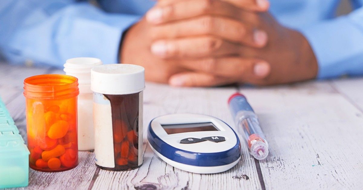 Person's hands folded near medication bottles, glucose meter, and insulin pen on a wooden surface.