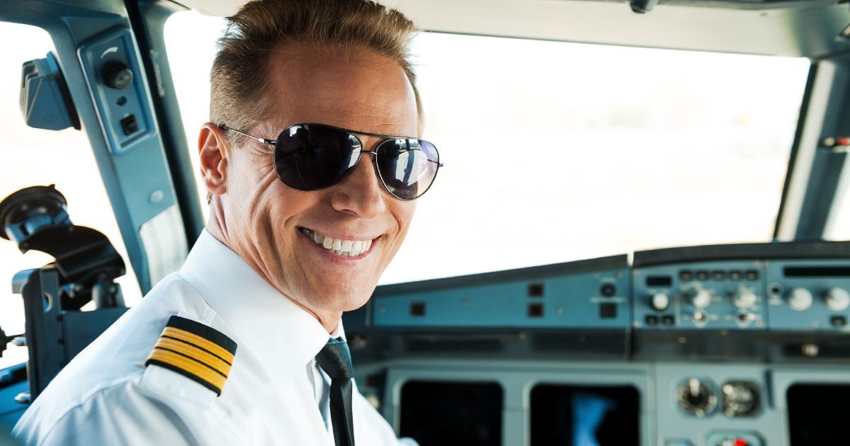 Pilot in cockpit smiling, wearing sunglasses and uniform.