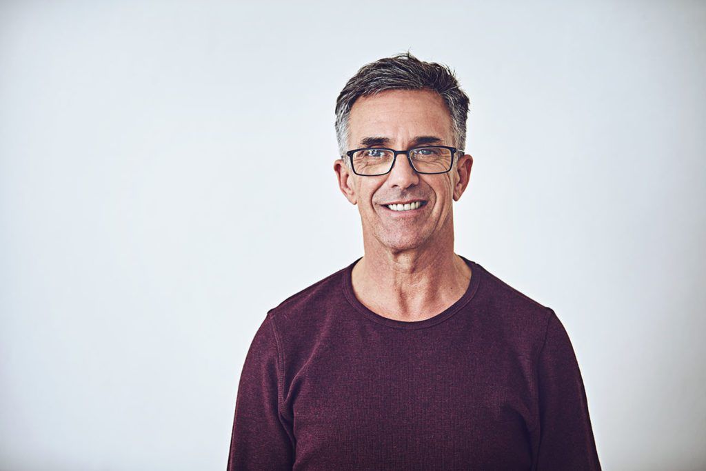 A man wearing glasses and a purple sweater is smiling for the camera.
