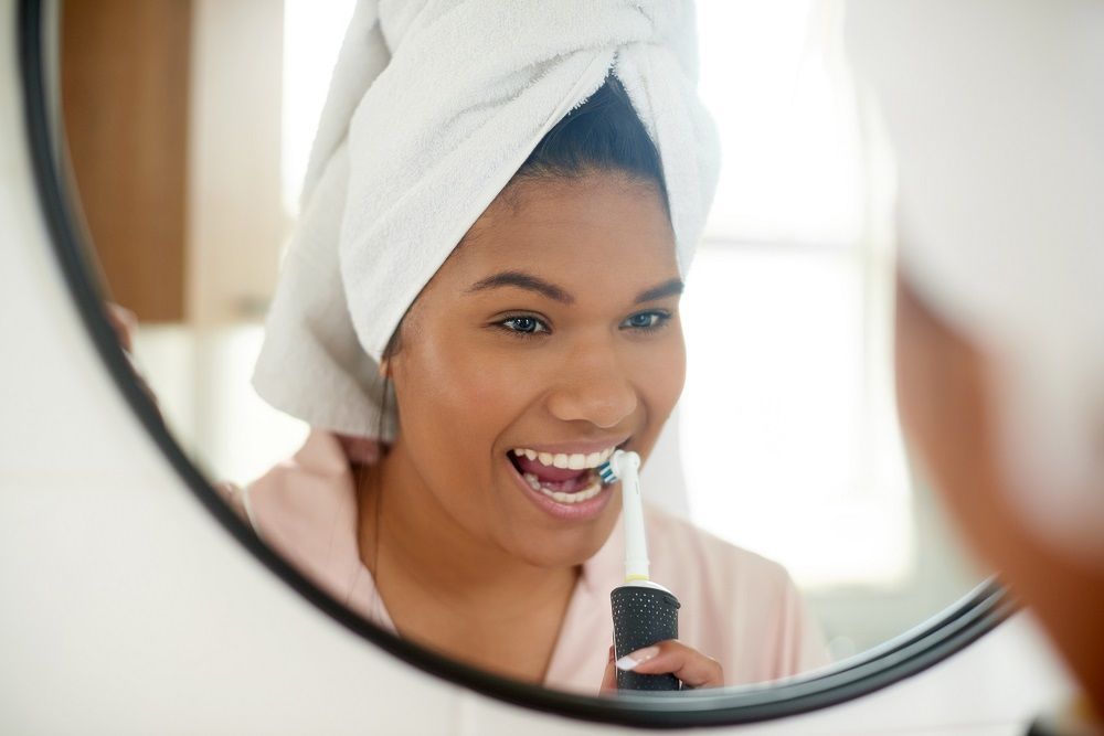 A woman with a towel wrapped around her head is brushing her teeth in front of a mirror.
