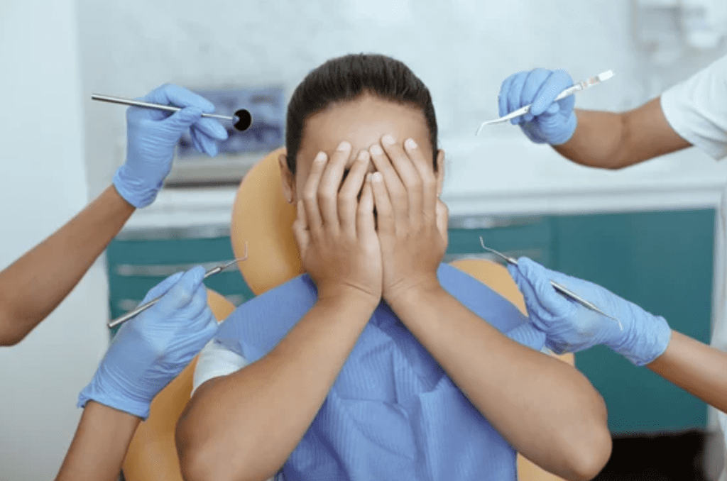 A woman is sitting in a dental chair covering her face with her hands.
