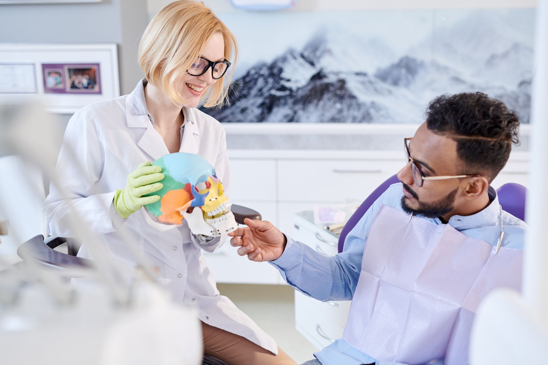 A dentist is talking to a patient in a dental chair while holding a model of a skull.