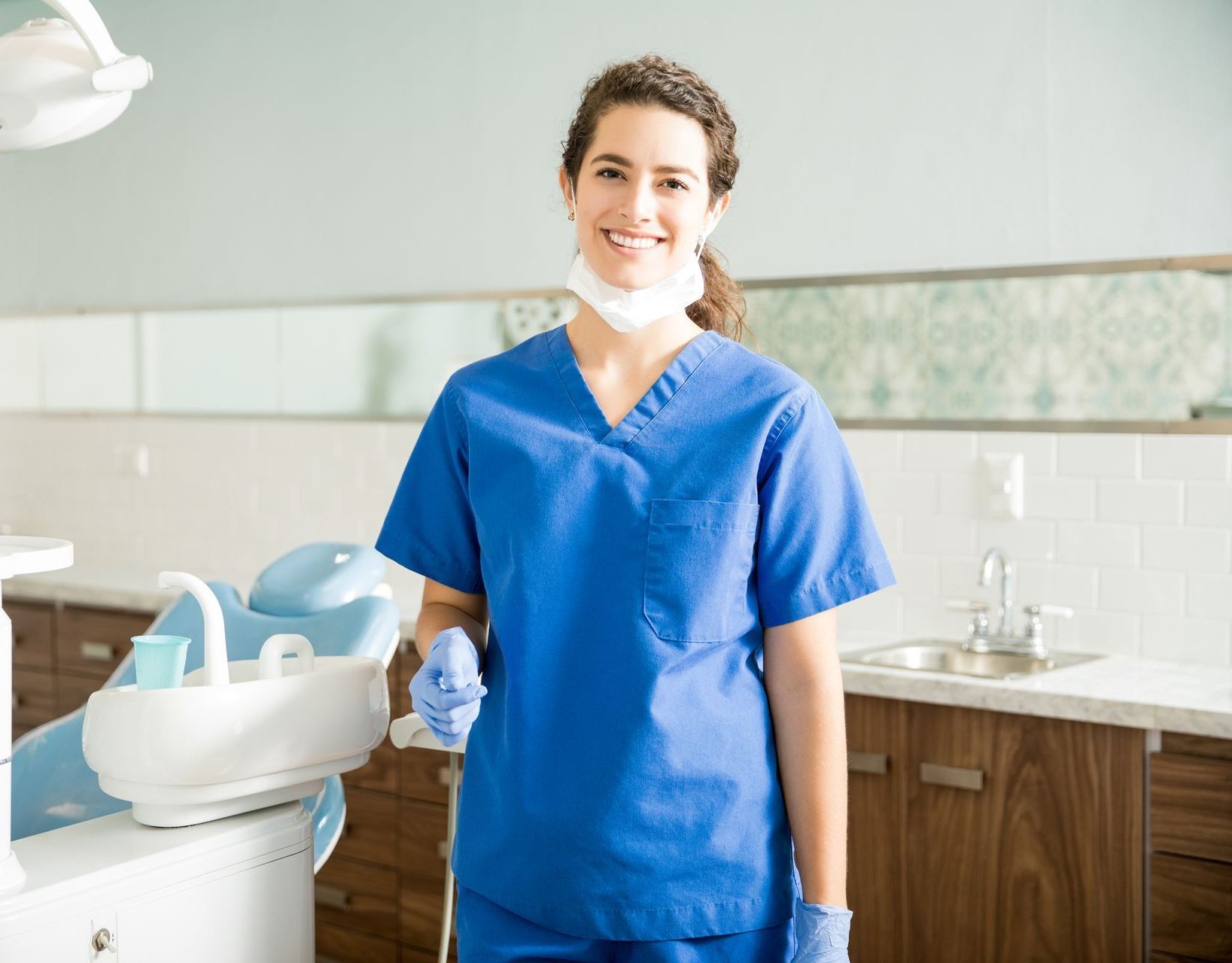 A female dentist wearing a mask and gloves is standing in a dental office.