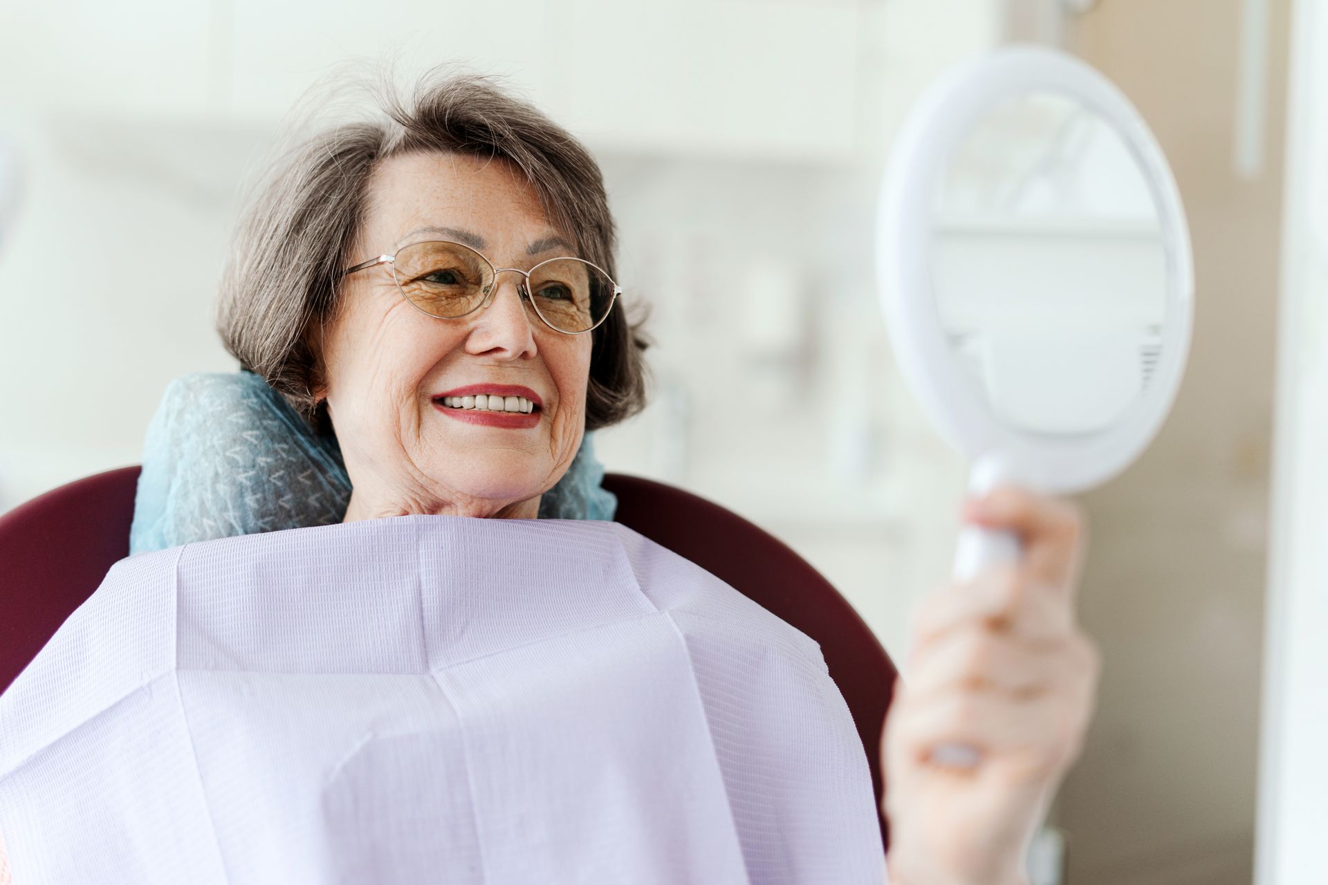 An elderly woman is sitting in a dental chair looking at her teeth in a mirror.