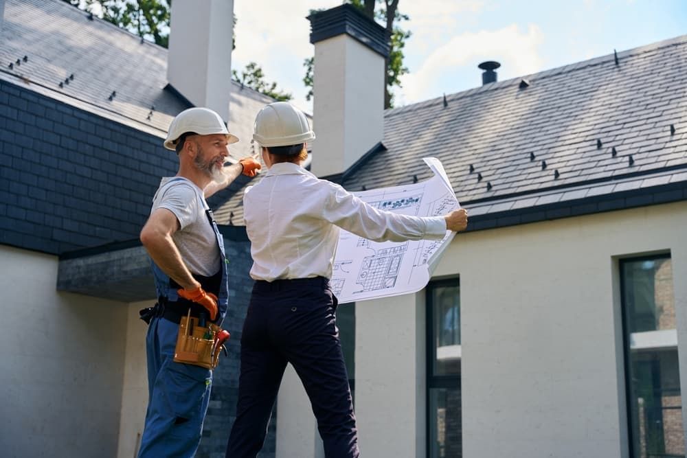 Two construction workers are standing in front of a house looking at a blueprint.