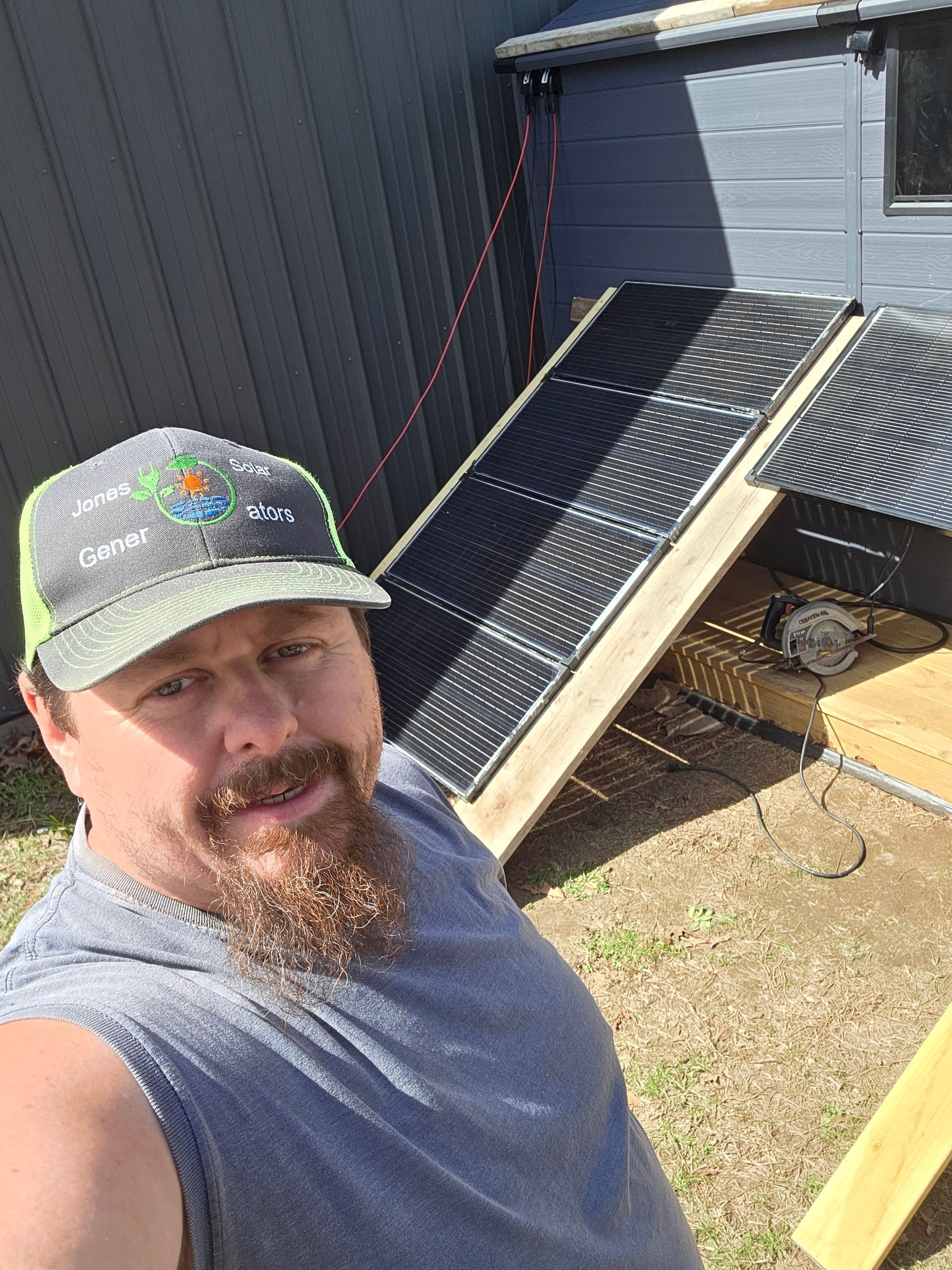 Man taking a selfie beside solar panels propped against a gray building in bright sunlight