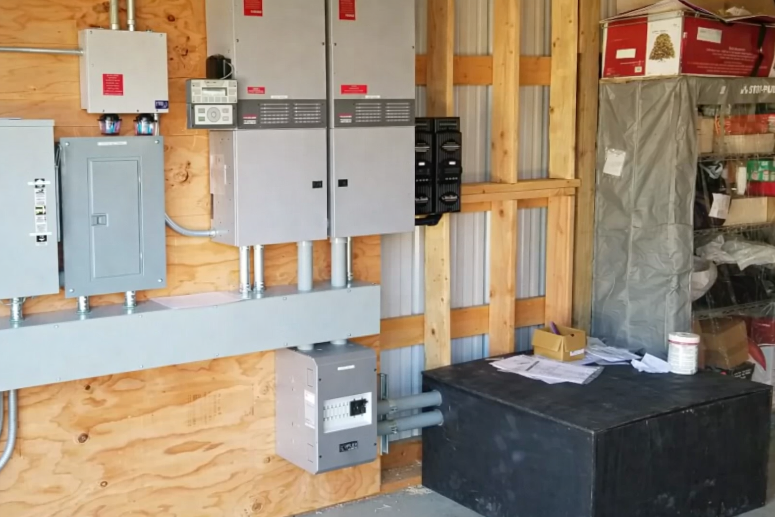 Solar power system with two inverters, electrical panels, and conduit mounted on a plywood wall in a utility area.