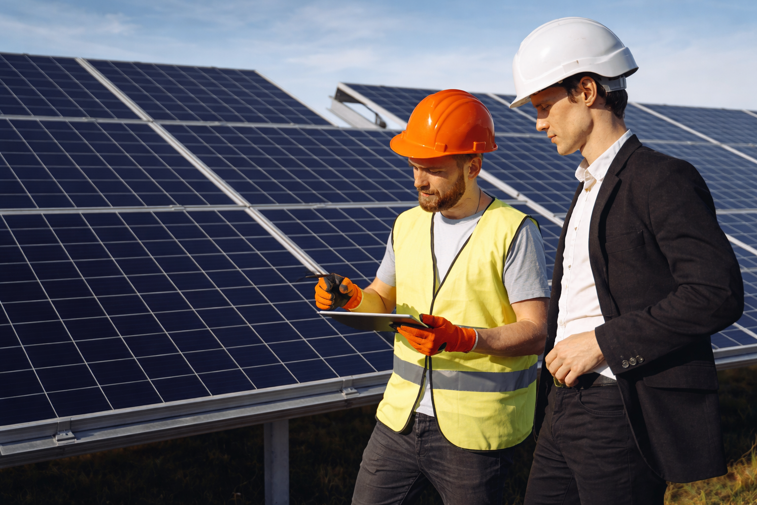 Two professionals in hard hats and work attire review data on a tablet in front of a large solar panel array.