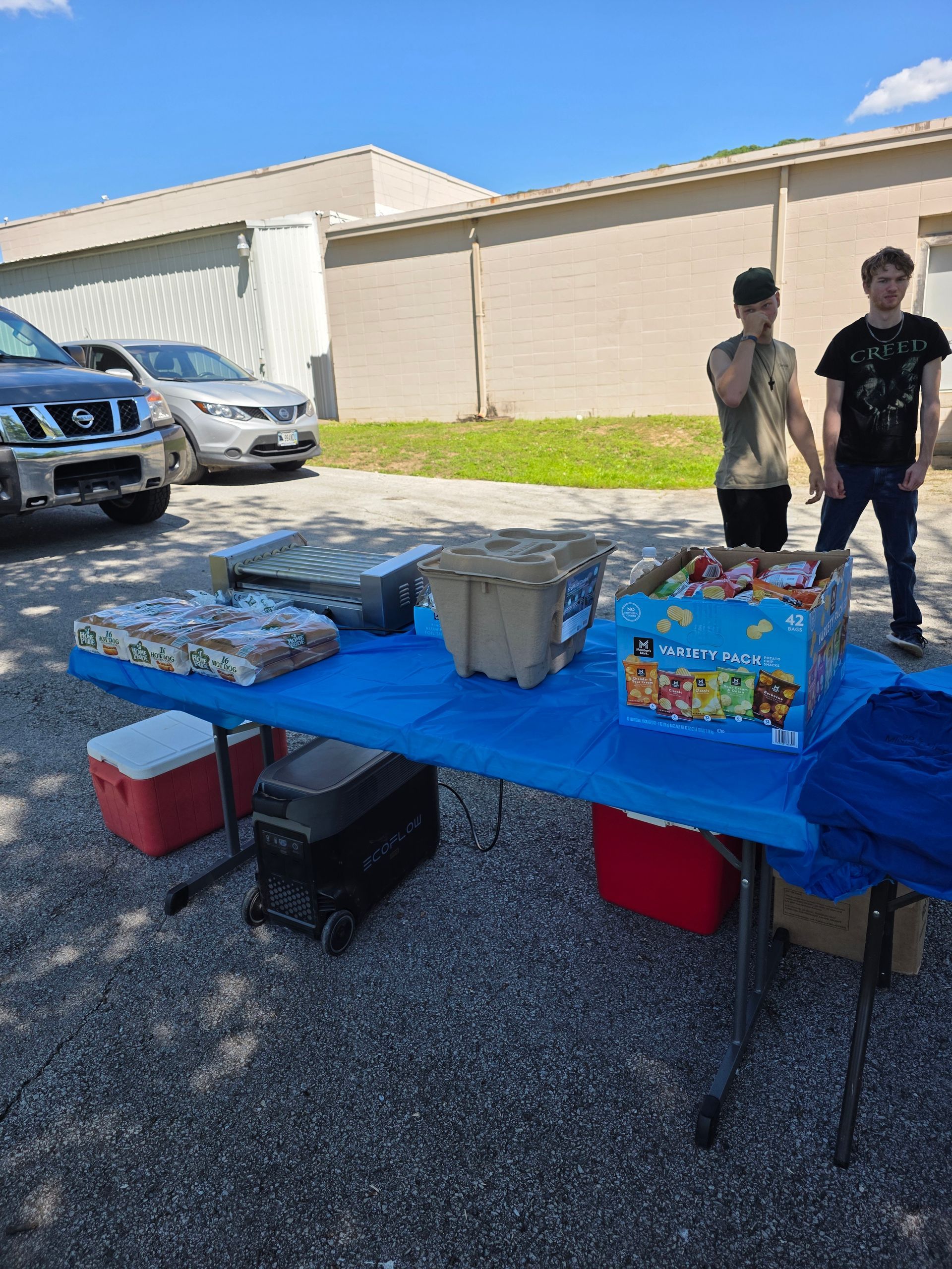 Blue-covered outdoor table with food containers; two people stand beside it near parked cars.