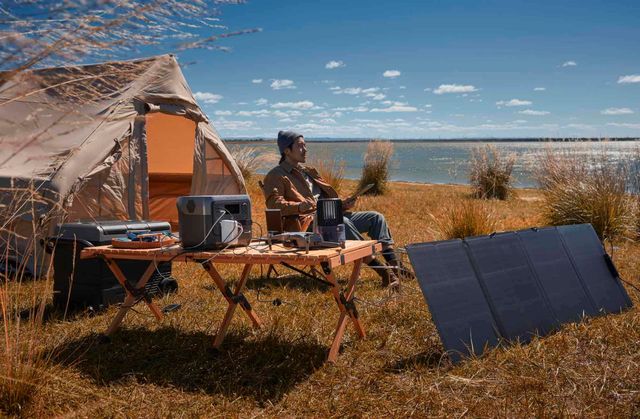 A solar panel charges a battery pack at a campsite, with tents and a person working on equipment in the background.
