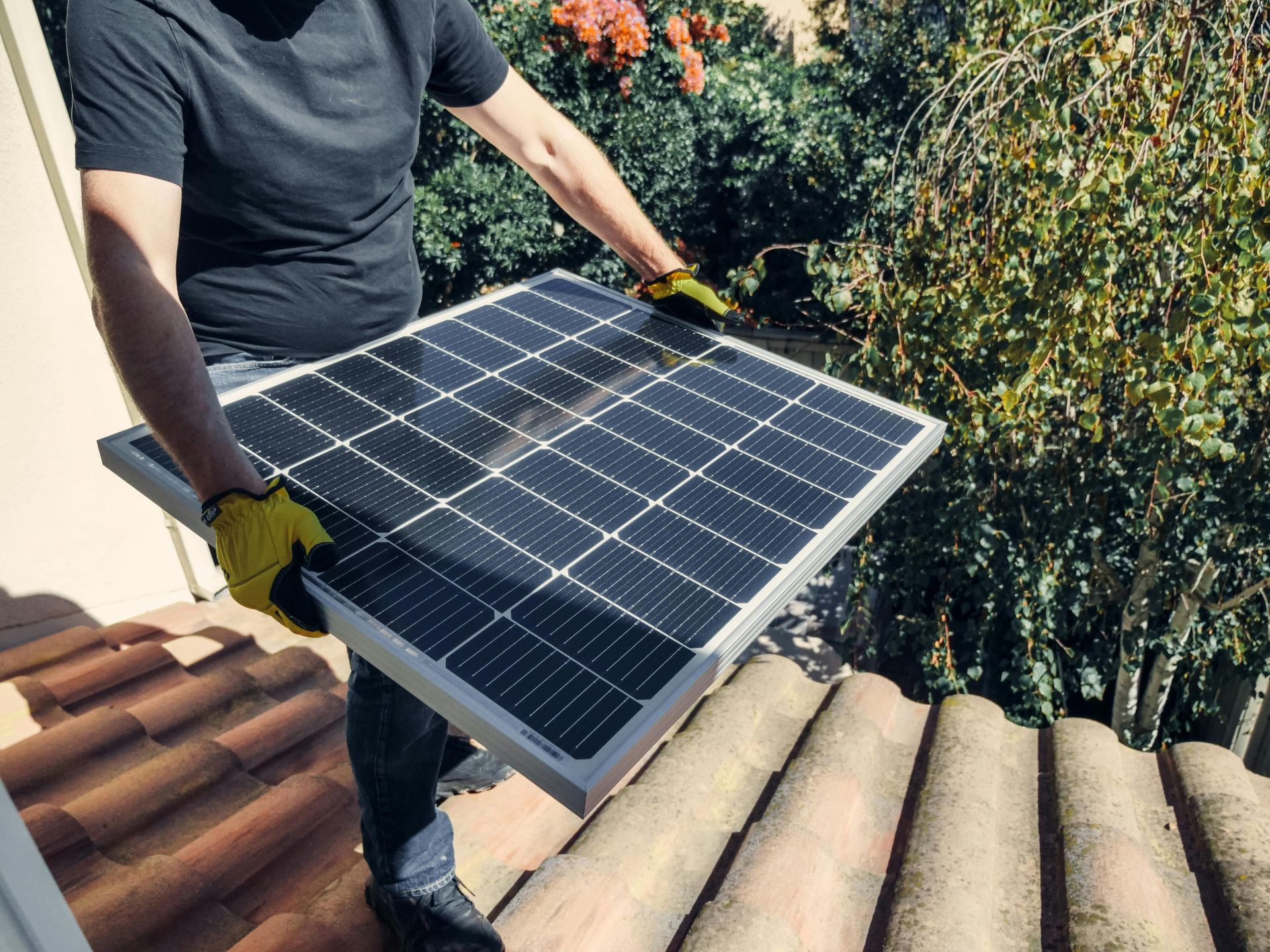 A person wearing work gloves holds a solar panel above a tiled roof.