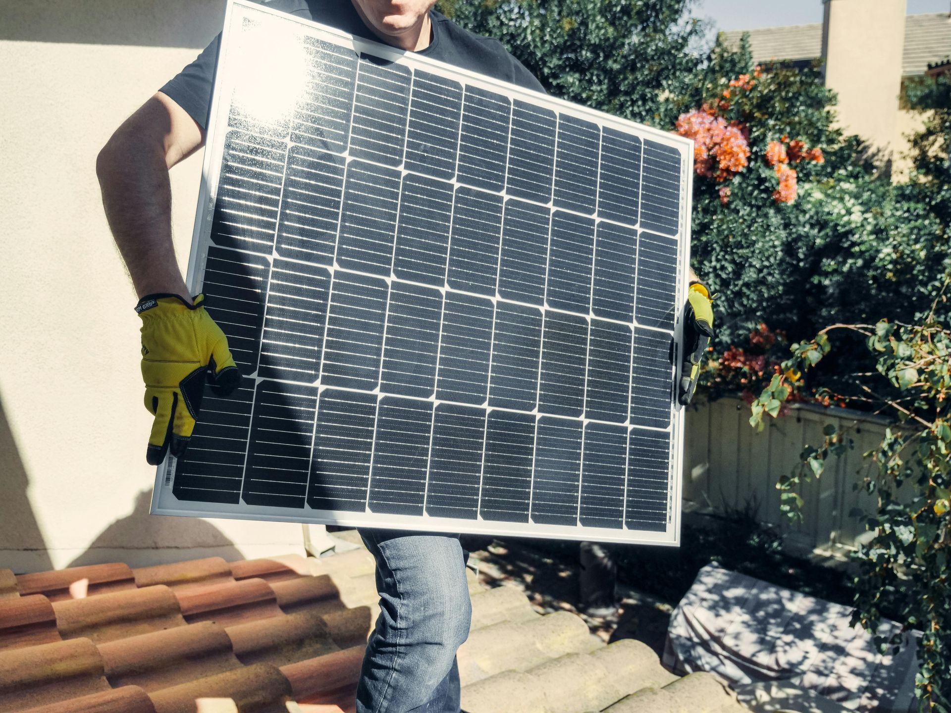 A person wearing yellow gloves holds a square solar panel in front of themselves on a tiled rooftop.