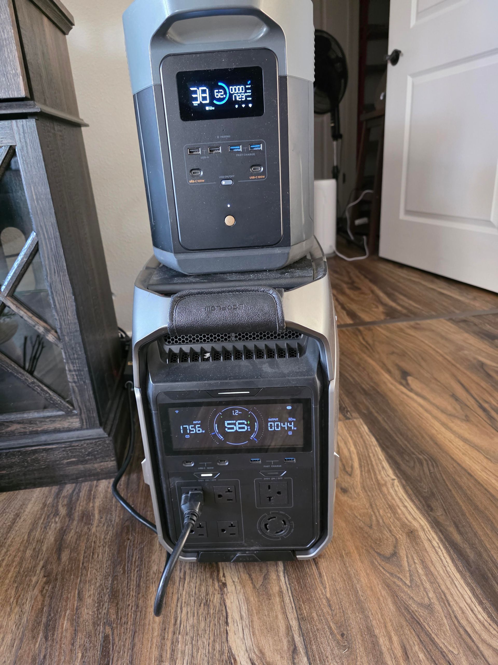 Two stacked black portable speakers with blue-lit displays on a hardwood floor near a white door.