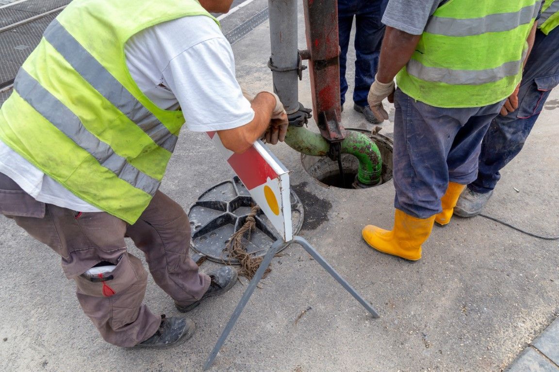 Workers in reflective vests inspect a manhole with tools on a sidewalk.