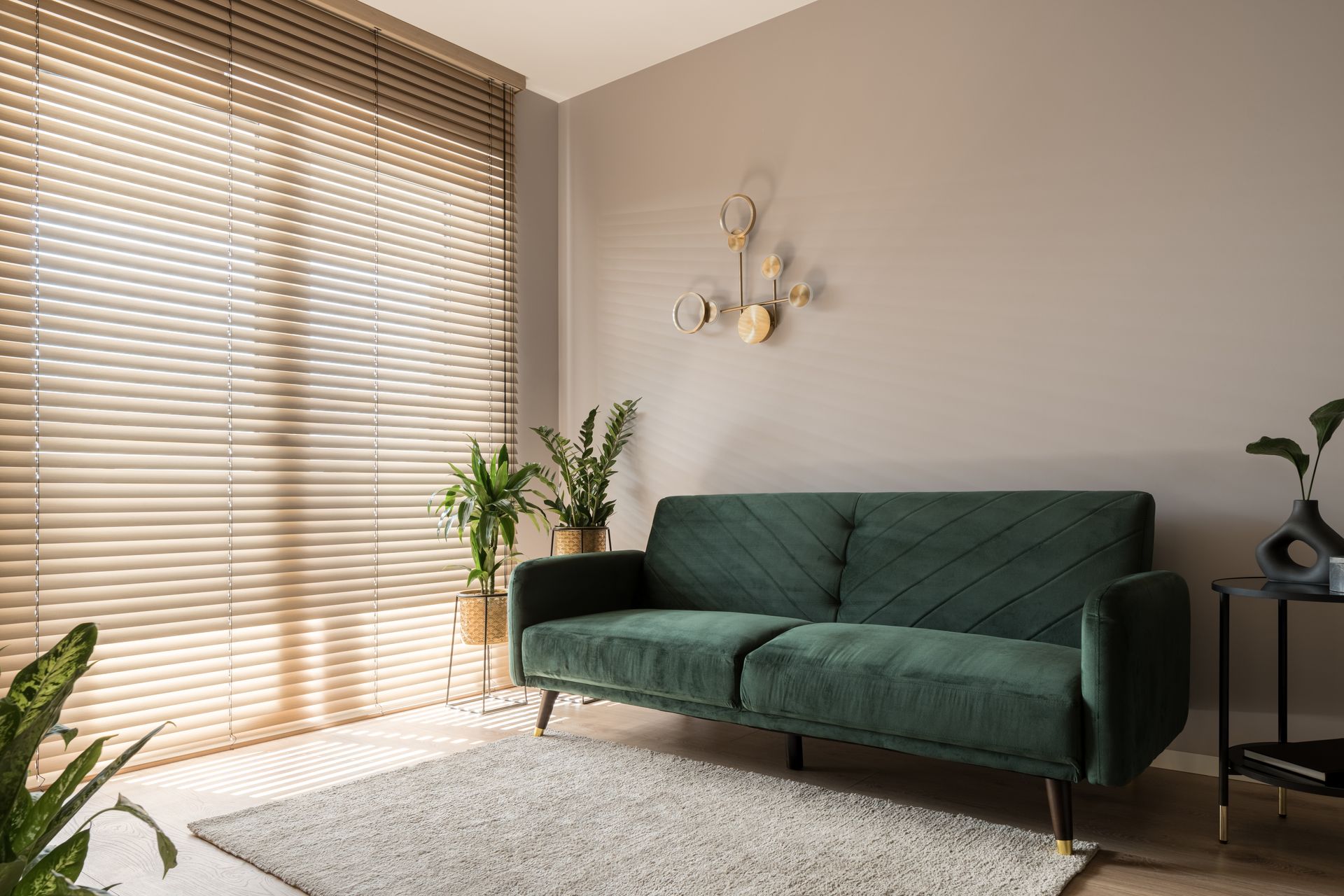 A neutral living room with house plants, a green couch, and gold-colored metal window blinds