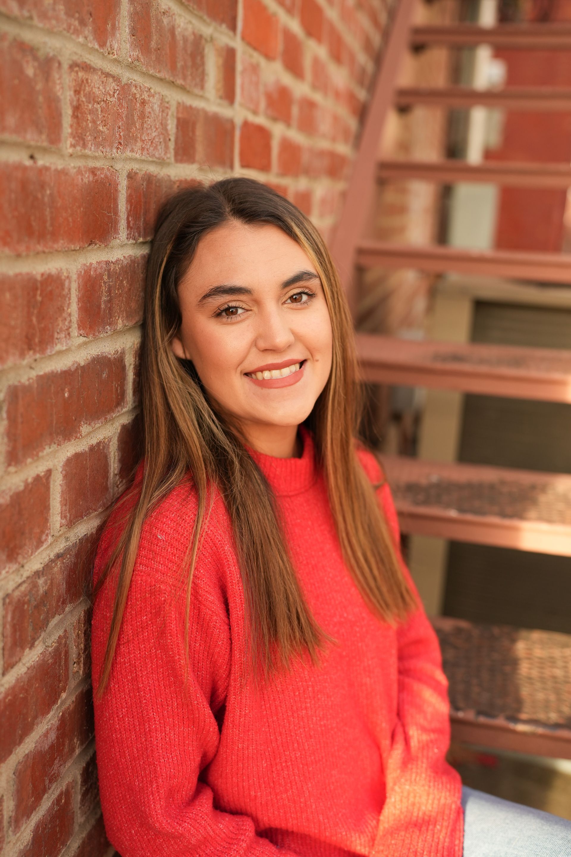 Woman in red sweater leans against brick wall, smiling. Metal stairs in the background.