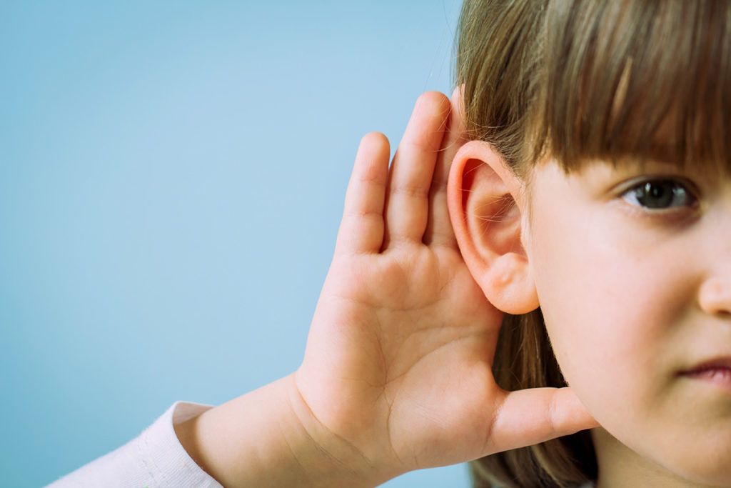 Young girl with her hand cupped behind her ear, listening. Light blue background.